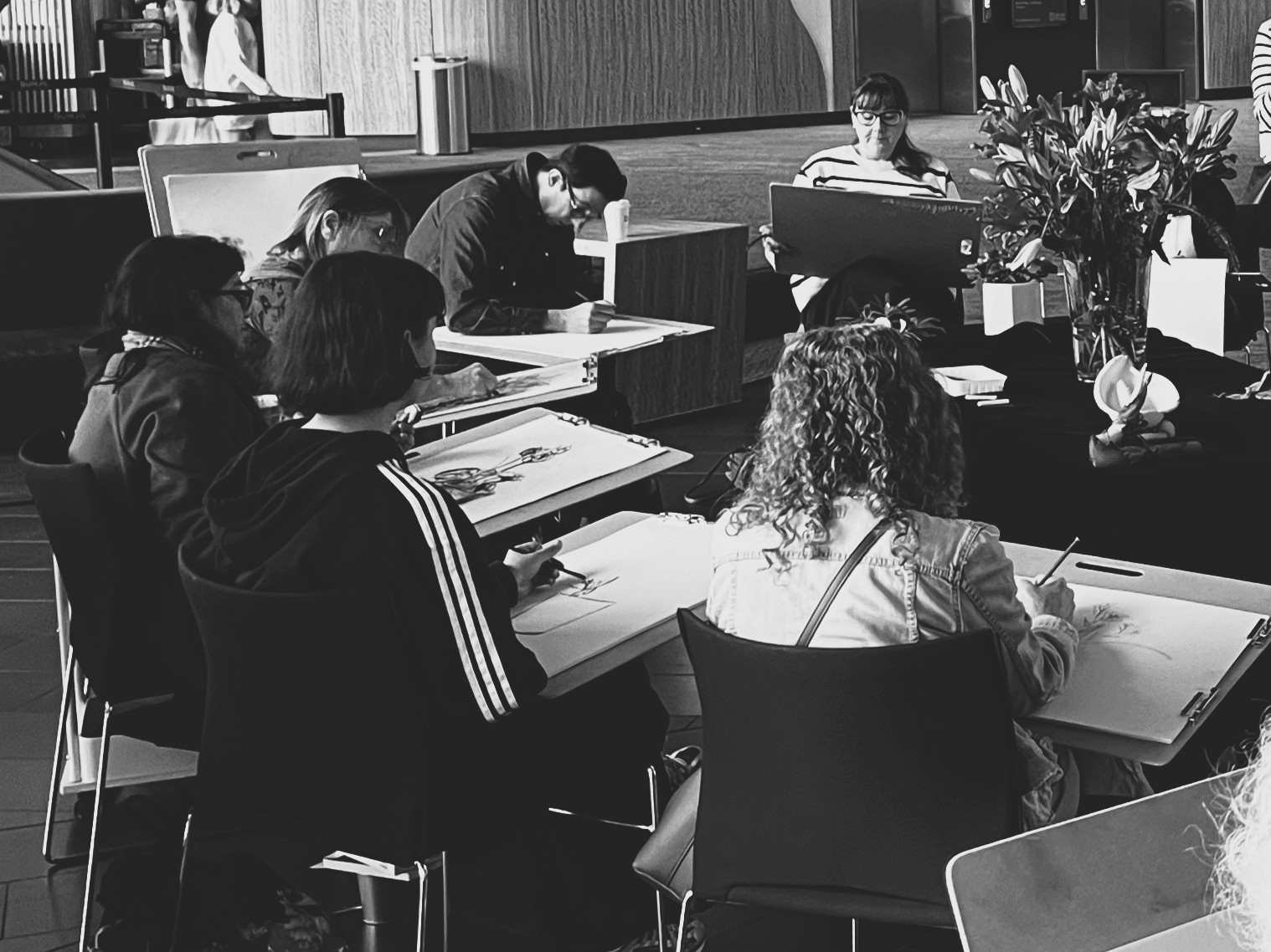 a group of people sitting on black chairs, sketching a still life display of flowers