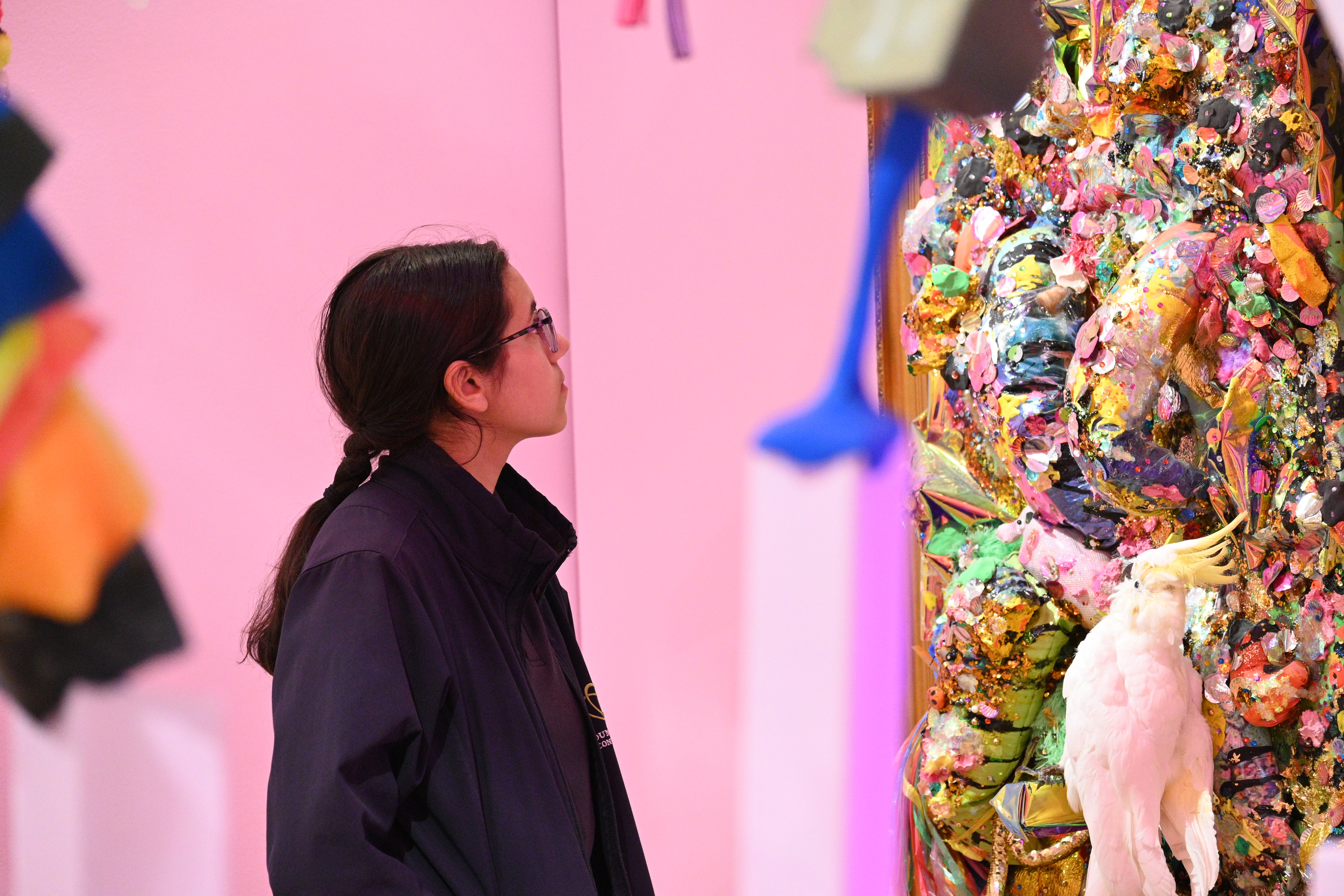 high school student wearing glasses and a navy blue uniform, looking at a multicoloured at installation with a pink background