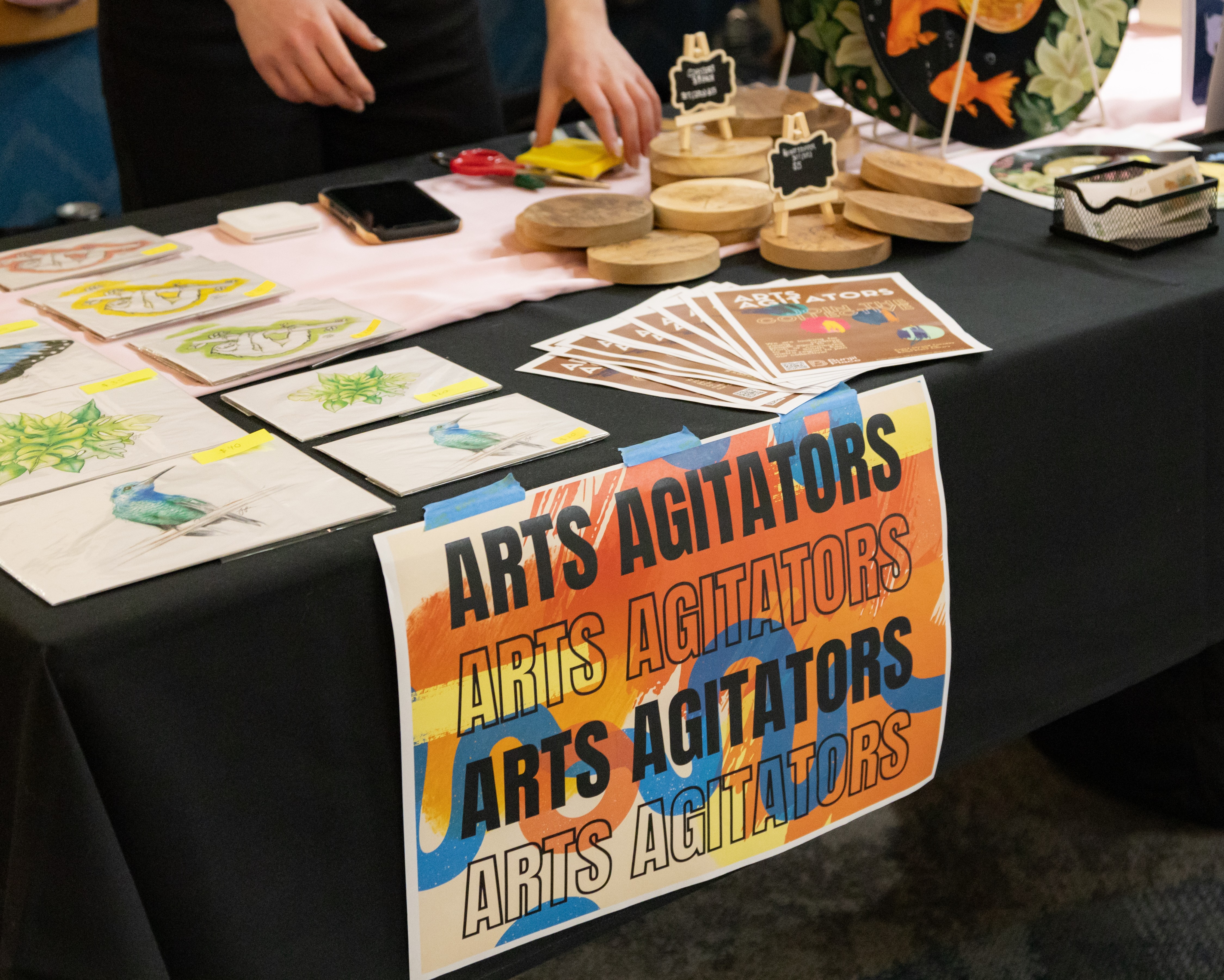 Image of signage that reads Arts Agitators. The sign has black writing on an orange background, and is taped to a black table cloth