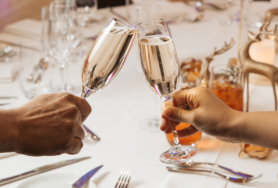 Two glasses of wine are held up, clinking together to mark "cheers" with the surrounding table set for a dinner