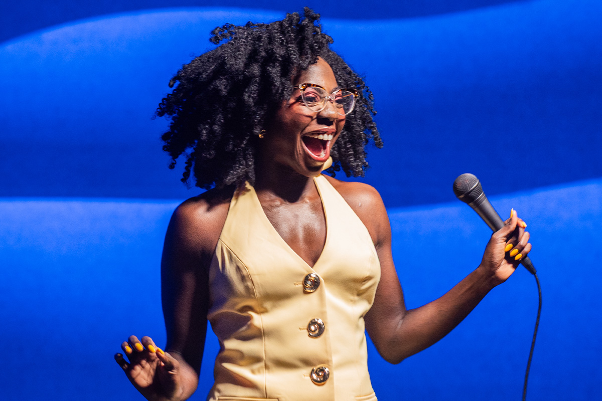 A woman in a yellow dress against a blue background, smiling as she holds a microphone - Anisa Nanduala