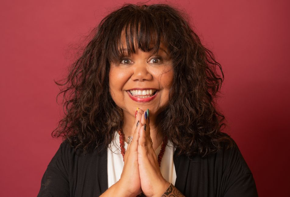 A woman with long curly hair holds her hands in front of her, as if praying, but she is smiling at the camera