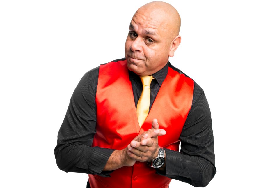 A bald man in a black shirt and red vest with a white tie looks sideways at the camera, 