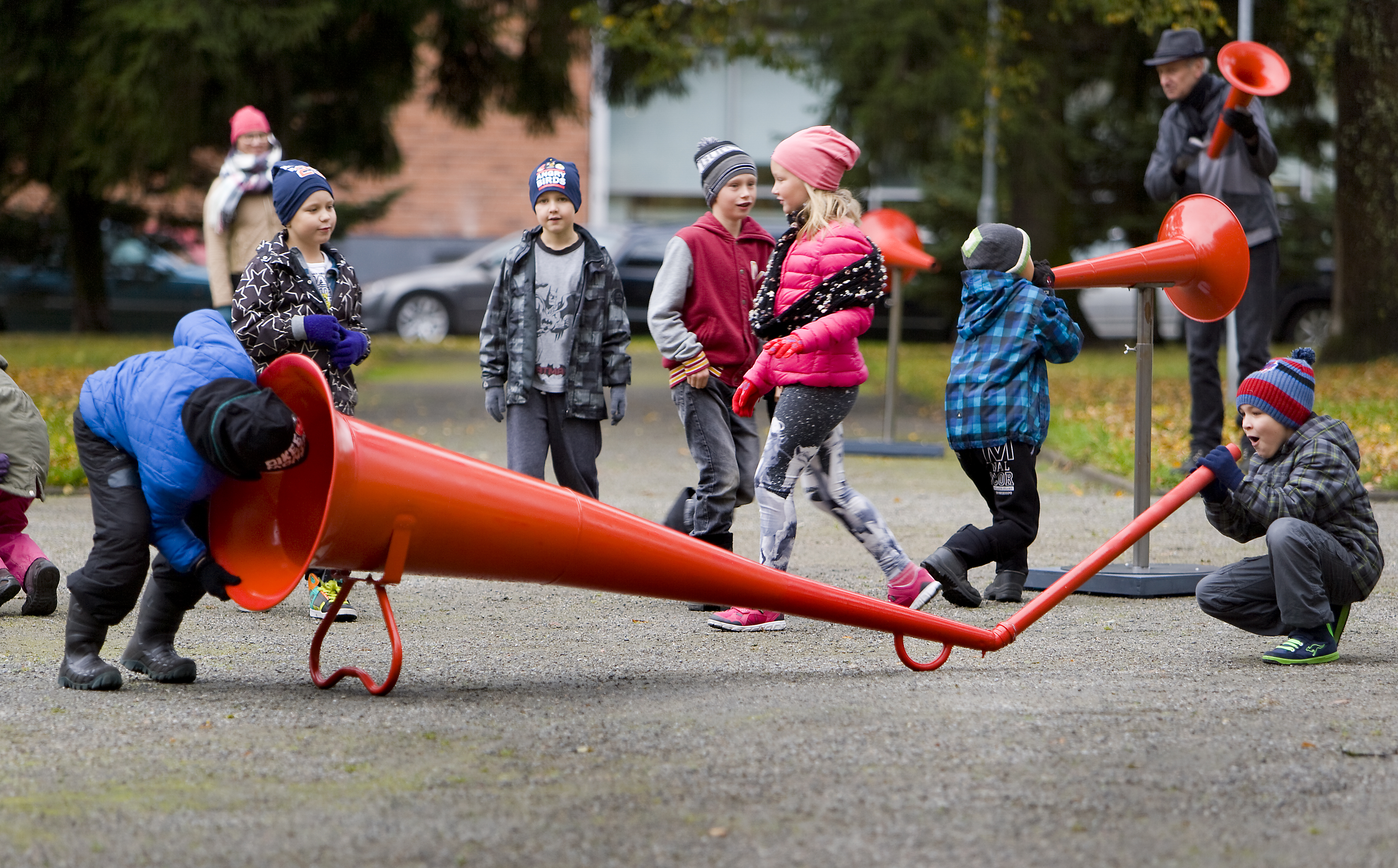Children in winter coats playing with a giant red megaphone in an outdoor park on a footpath. One is speaking into one end of the megaphone, while the other listens at the other end. while
