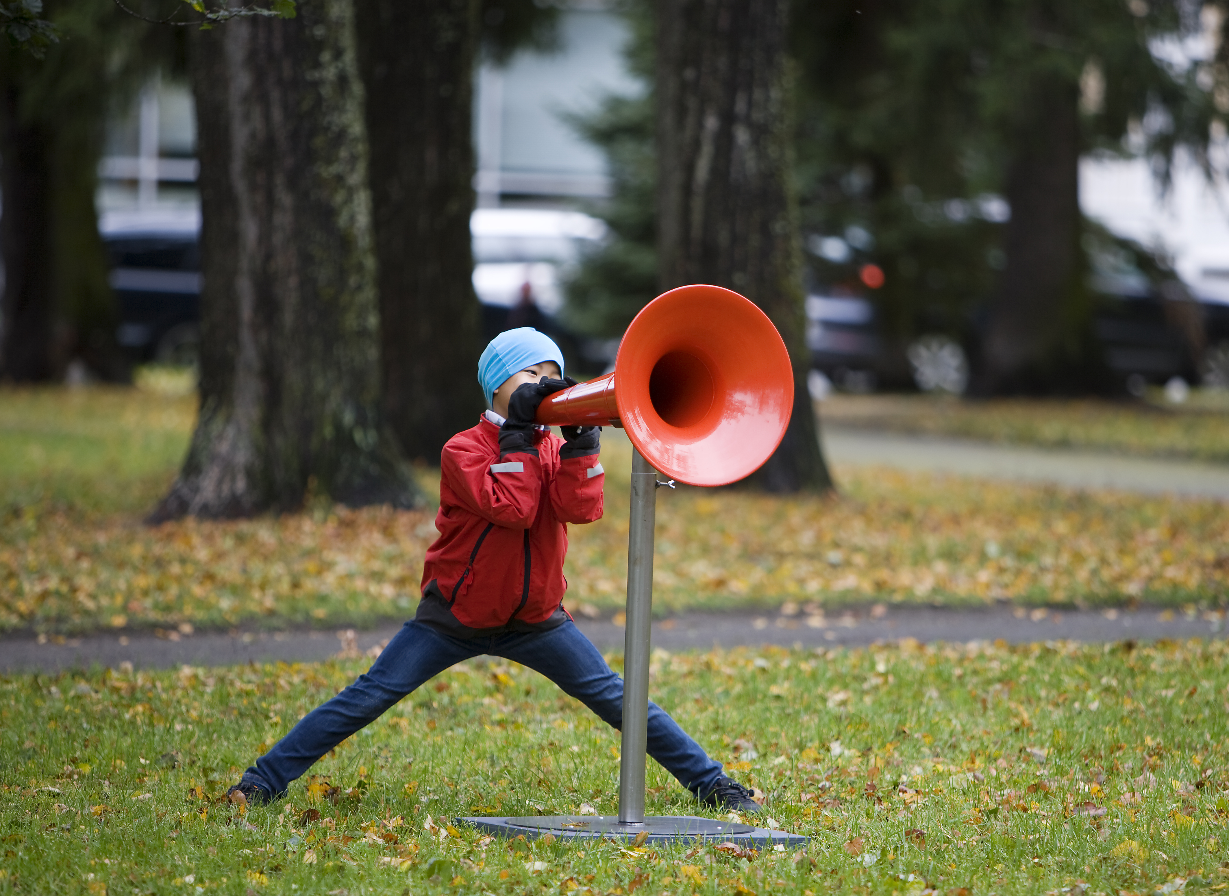 A young boy is speaking into a large red megaphone on a stand. He is wearing a winter coat and beanie, and is in a park.