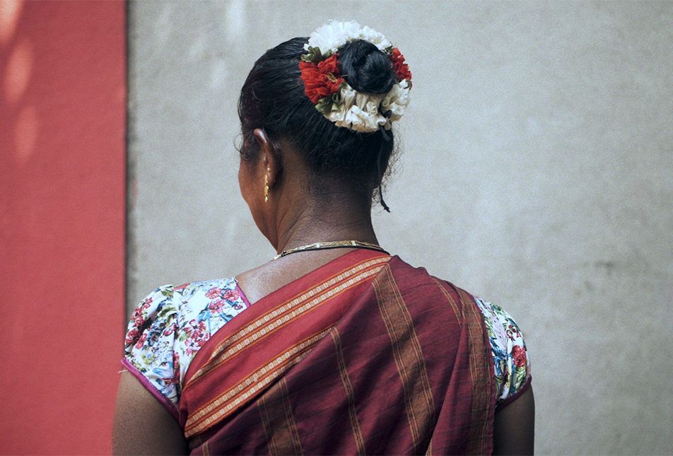 A woman facing the wall, wearing a sari and floral details in her hair