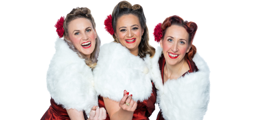 Three women with white shawls and red rosettes in their hair and bright red lipstick smile at the camera. 