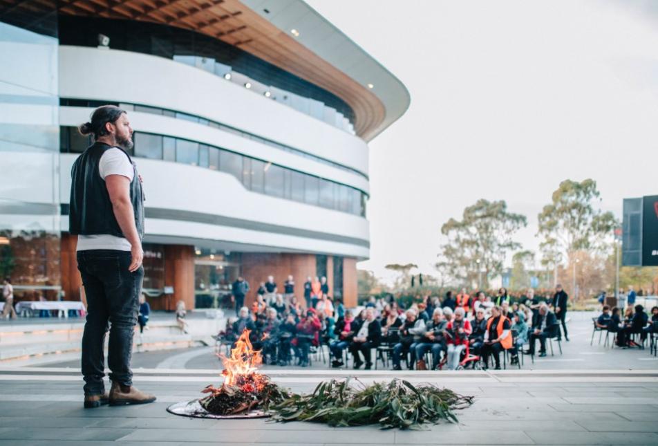 A man stands in front of a fire outside a building, as part of a smoking ceremony