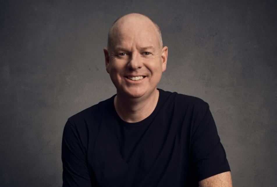 Tom Gleeson - a comedian, sits smiling in front of a grey background