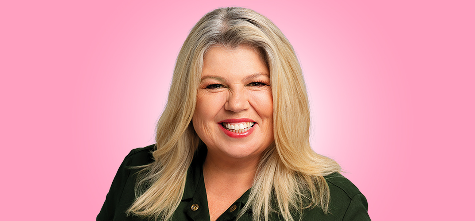 Urzila Carlson, a female comedian with blonde hair, stands in front of a pink background.
