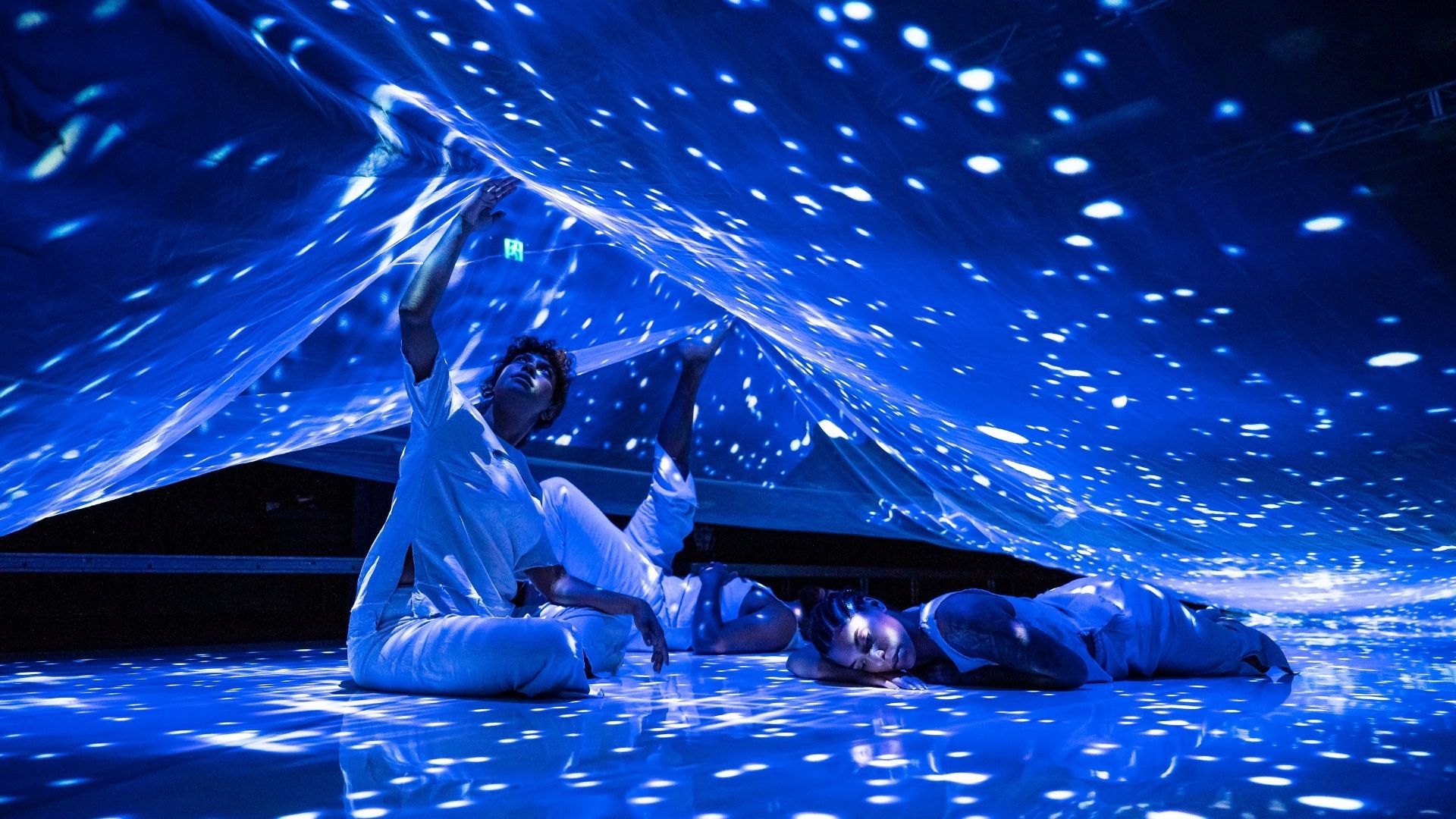 Two people holding a shimmery veil in a dark room, wearing white clothes, sitting on the floor. Another person lies underneath