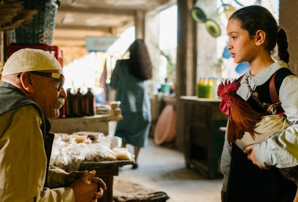 A young girl holding a rooster talking to an older, seated man in a market.