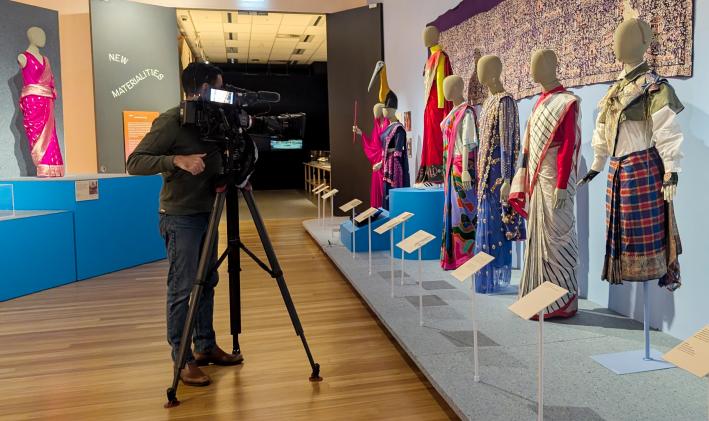 A camera man with a mounted TV camera pointed at a row of mannequins wearing saris in the Bunjil Place Gallery 'The Offbeat Sari' exhibition.