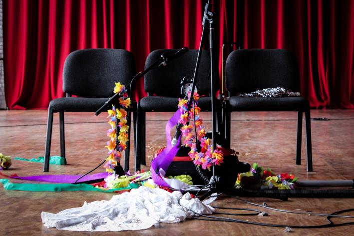 Three black folding chiars in front of a red Theatre curtain, with various microphones and flowers draped all over them.