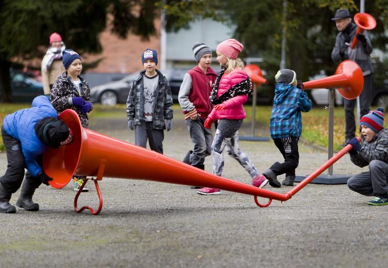 Children in winter coats playing with a giant red megaphone in an outdoor park on a footpath. One is speaking into one end of the megaphone, while the other listens at the other end. while