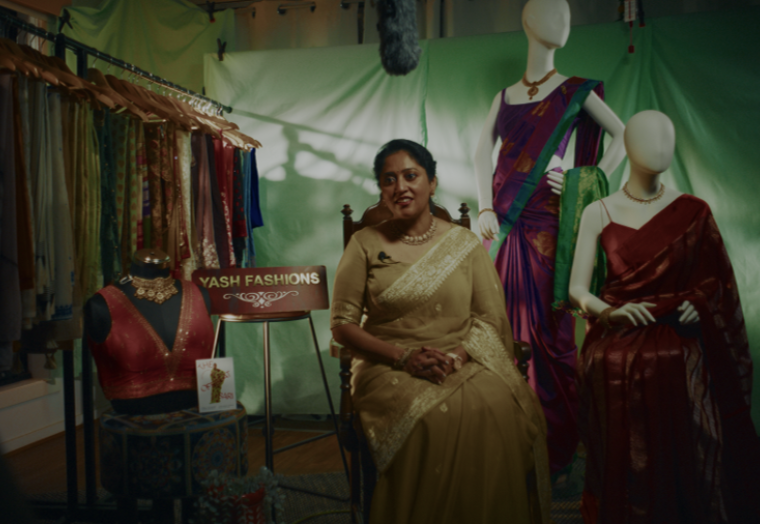 a woman in a yello sari sitting next to a display of many colourful saris