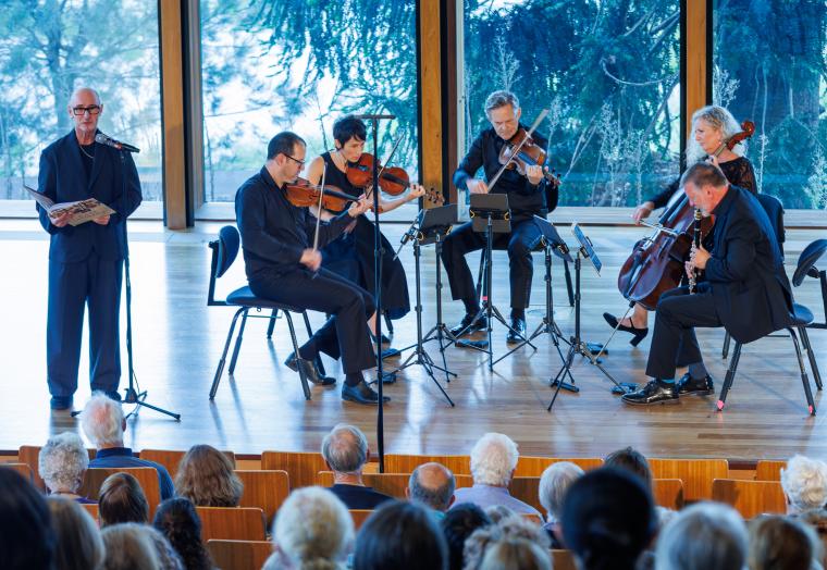 Five seated musicians on a stage performing various string instruments, with a man standing in front of a microphone holding a large book