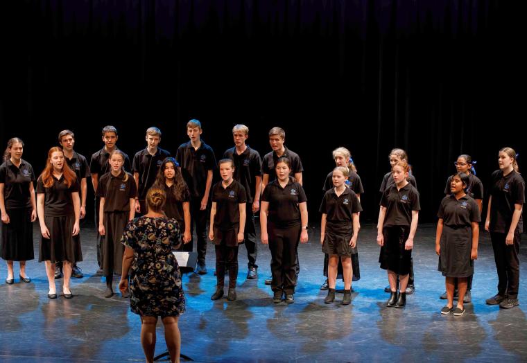 A group of teenagers/youths wearing black singing on a stage, with a female conductor facing towards them.