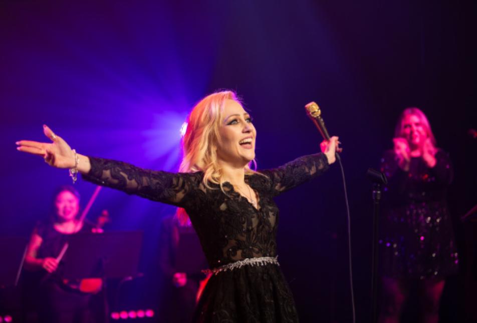 A blonder woman in a black dress holds her arms out on stage - To Be Loved - A Dedication to Adele