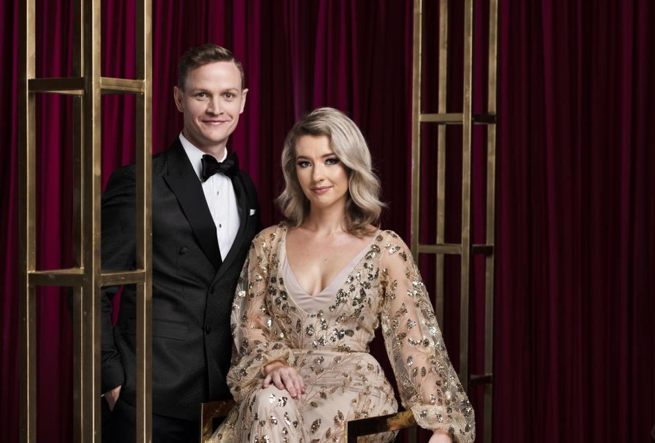 A man in a suit and a woman in a gold glittery dress look seriously at the camera