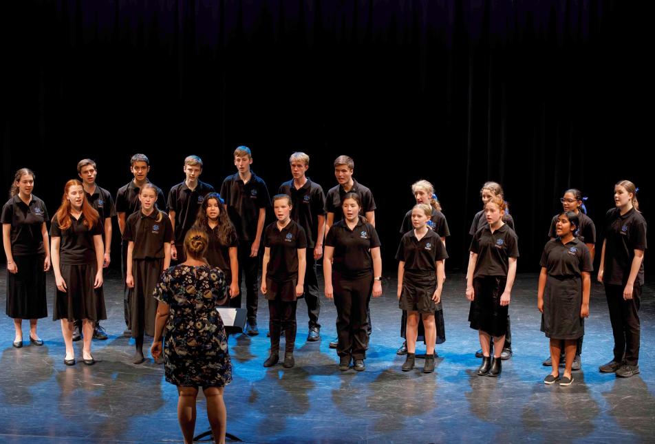 A group of teenagers/youths wearing black singing on a stage, with a female conductor facing towards them.