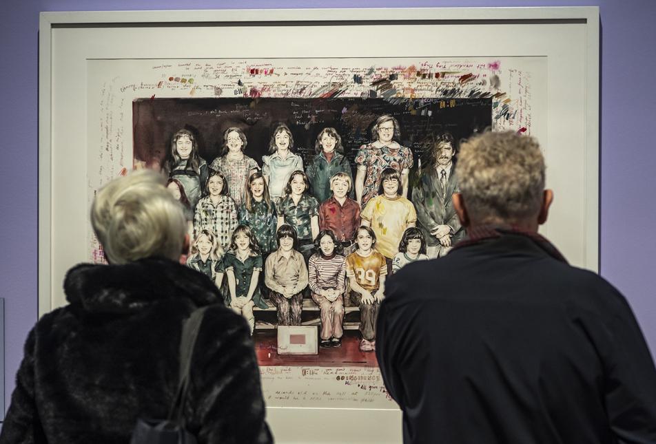 Two people stand with their back to the camera looking at an artwork on the wall - a painting of a class photograph