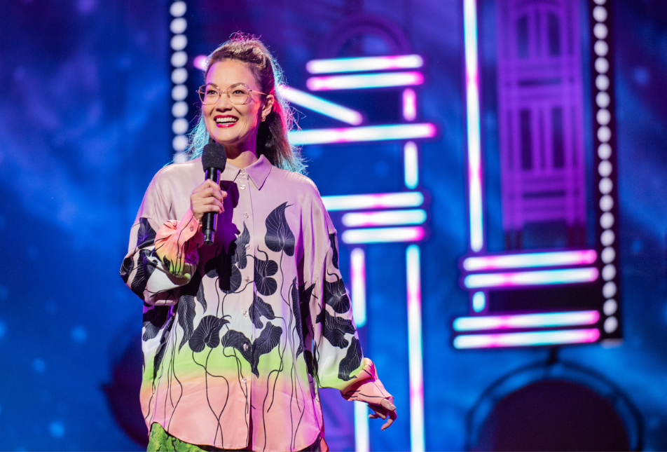 A woman in a colourful top, wearing glasses, on stage. 