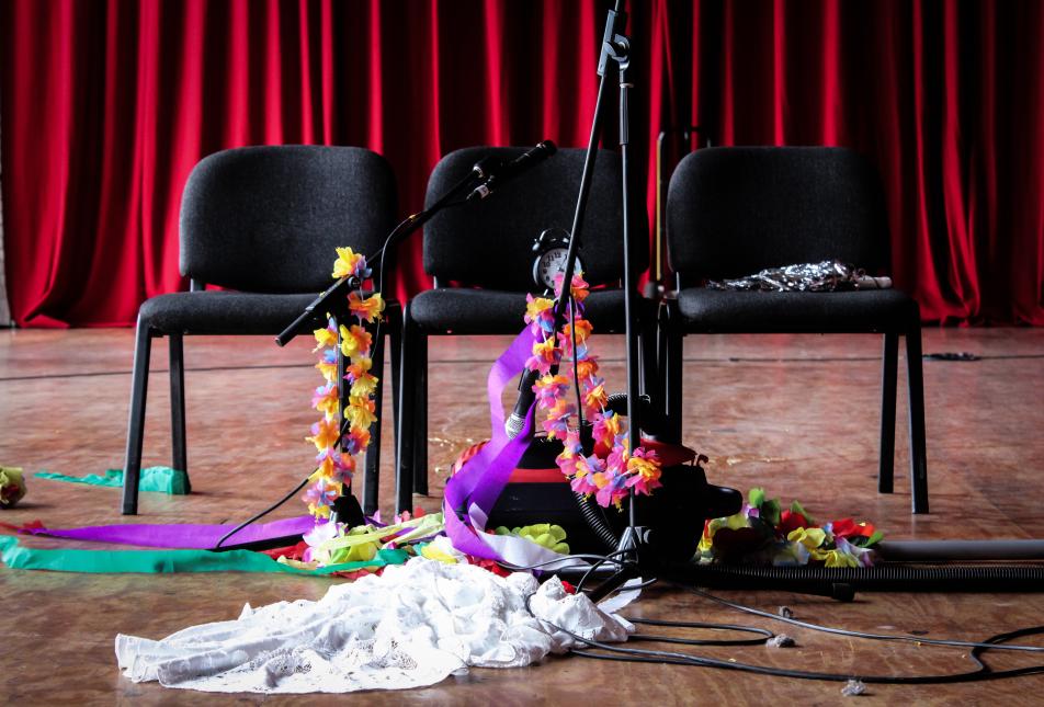 Three black folding chairs sitting in front of a red Theatre curtain, with various microphone stands decorated with flowers and other costume accessories.