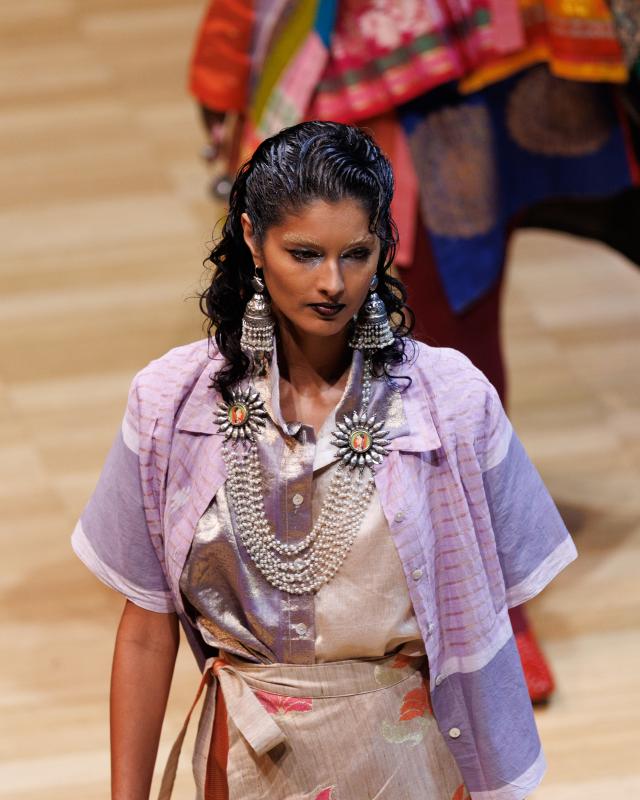 A person walking on a runway wearing a lilac coloured shirt and skirt and large silver earrings