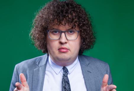 A man in a grey suit with curly hair and glasses points at the camera_Ray O'Leary
