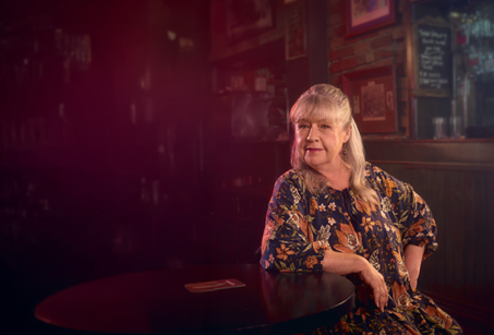 A woman leans on a table, with a bar in the background