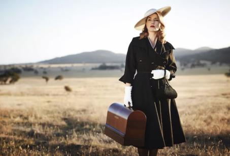 A film still showing a women in a nice dress and hat, holding a leather bag, standing in a rural setting, like an open field. 