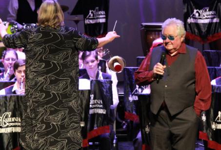 Image of an orchestra in action with a man in a red shirt and black vest ready to talk into a microphone