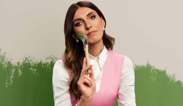 A woman with long brown hair in a white shirt and pink vest holds a paintbrush against her face - comedian Melanie Bracewell
