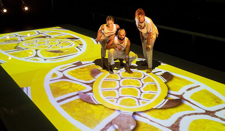Three indigenous performers stand on an art digital projection