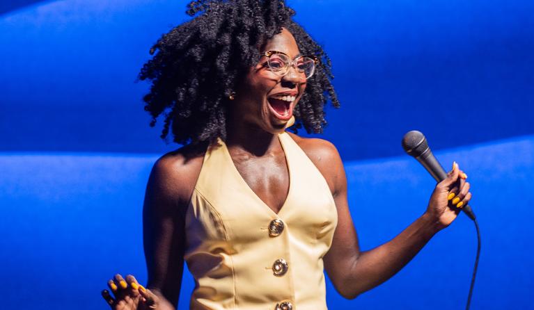 A woman in a yellow dress against a blue background, smiling as she holds a microphone - Anisa Nanduala