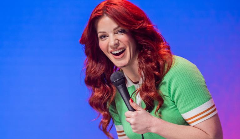 A woman with red hair, wearing a green dress, holds a microphone - Bron Lewis