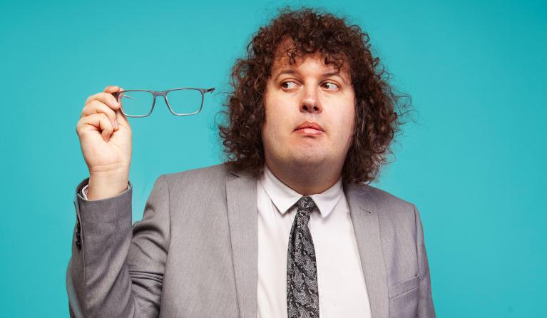 A man in a grey suit holds his glasses to one side - comedian Ray O'Leary
