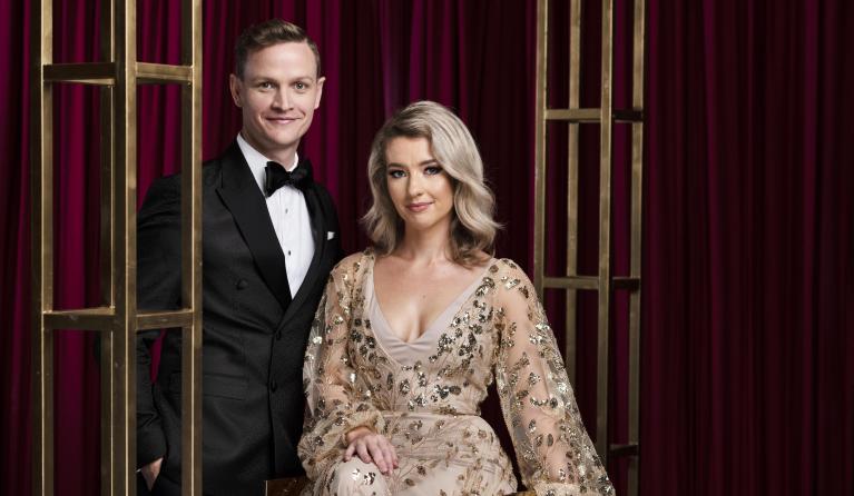 A man in a suit and a woman in a gold glittery dress look seriously at the camera