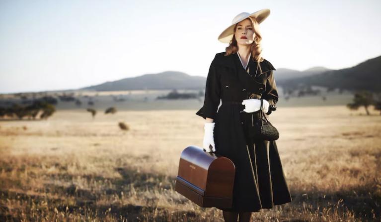 A film still showing a women in a nice dress and hat, holding a leather bag, standing in a rural setting, like an open field. 