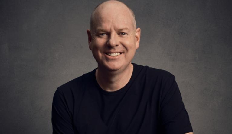 A headshot of a bald man in a black t-shirt grinning at the camera