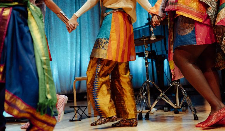 Three people stand in a circle holding hands, shown from the waist down. They wear brightly coloured, patterned garments made from saris and distinct shoes, with a drum kit and blue curtain visible in the background, suggesting a live performance setting.