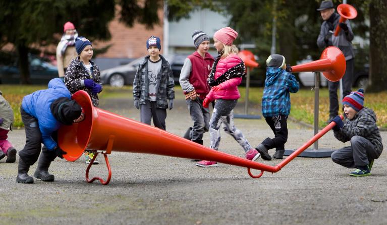 Children in winter coats playing with a giant red megaphone in an outdoor park on a footpath. One is speaking into one end of the megaphone, while the other listens at the other end. while