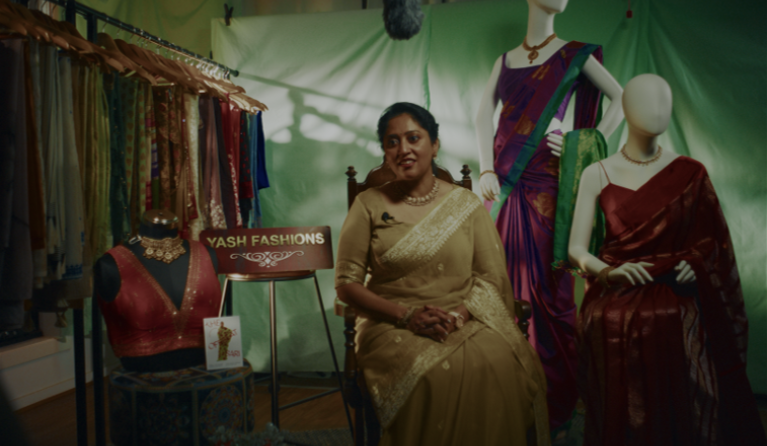 a woman in a yello sari sitting next to a display of many colourful saris