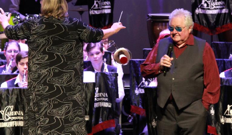Image of an orchestra in action with a man in a red shirt and black vest ready to talk into a microphone