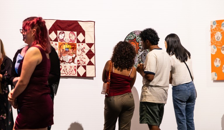 three people looking closely at an artwork on a wall. the artwork is obscured from view. next to the group of people is a white and red quilted artwork