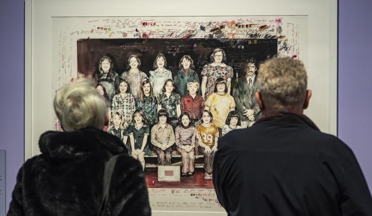 Two people stand with their back to the camera looking at an artwork on the wall - a painting of a class photograph