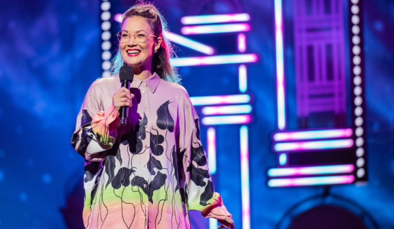 A woman in a colourful top, wearing glasses, on stage. 