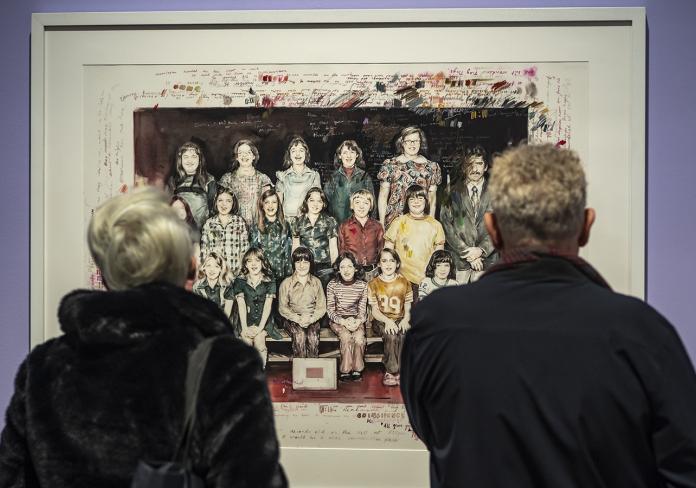 Two people stand with their back to the camera looking at an artwork on the wall - a painting of a class photograph