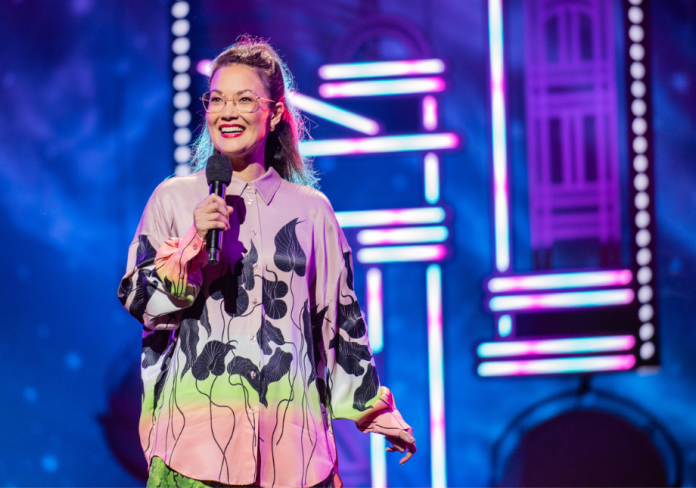 A woman in a colourful top, wearing glasses, on stage. 